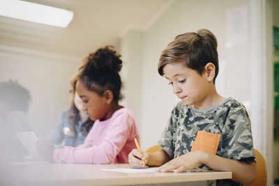 Boy sitting with friend writing on paper at desk in classroom