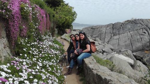 Portrait of friends sitting on retaining wall by plants