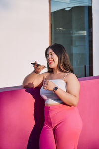 Glad plus size female in stylish clothes recording audio message via smartphone and smiling while leaning on pink wall of modern building near window on sunny day on city street