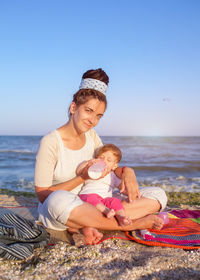 Portrait of smiling mother feeding milk to daughter while sitting at beach during sunset