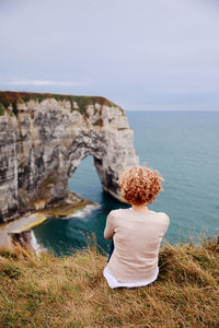 Rear view of woman sitting at cliff by sea against sky