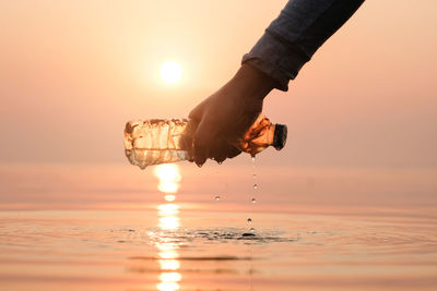 Reflection of hand holding sun in sea against sky during sunset