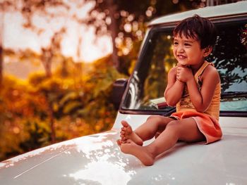 Full length of cute smiling girl looking away while sitting on vehicle hood during sunset