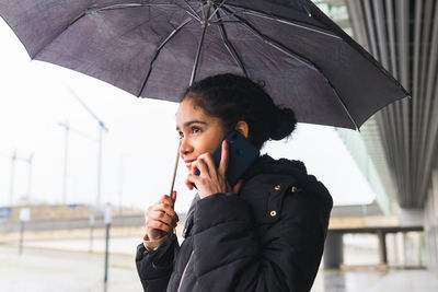 Cheerful colombian female in outerwear standing with umbrella on a phone call on mobile phone at street