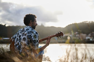 Back view of male musician playing acoustic guitar while sitting on seashore on sunny day