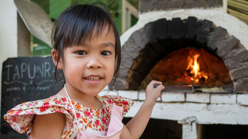 Little girl makes hand made pizza and stands smiling in front of a wooden brick pizza oven.