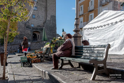 Rear view of people sitting on bench against buildings in city