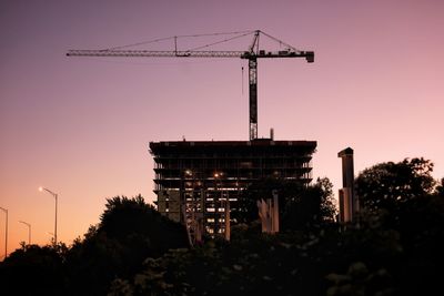 Low angle view of silhouette crane by building against sky during sunset