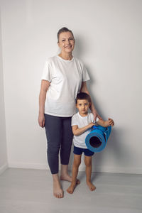 Elderly female athlete with a baby boy is standing at home with a yoga mat