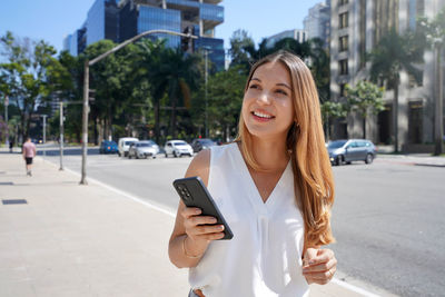 Beautiful young woman holding smartphone walking in financial district. 