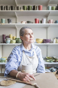 Senior female potter looking away while sitting at table with vase in workshop