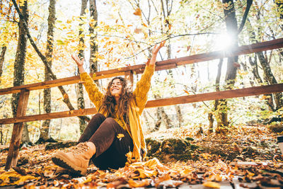 Positive curly haired woman in yellow sweater throwing dry leaves in picturesque autumn forest 