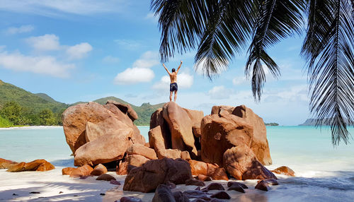 Young man with outstretched arms on rocks on sandy tropical beach.