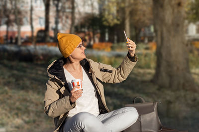 Young woman with a cup of drink and smartphone sits cross-legged on bench on a sunny day. soft focus