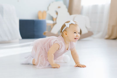 A pretty blue eyed blonde one year old in a pink dress crawls on all fours in the nursery