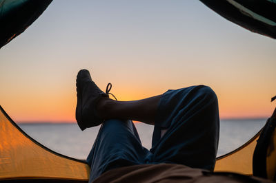 Low section of man relaxing in tent by sea against sky during sunset