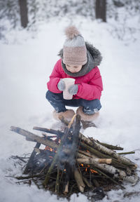 Cute girl drinking hot chocolate while crouching by campfire during winter