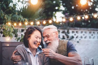 Man feeding woman against illuminated lighting equipment and trees