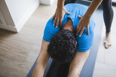 High angle of unrecognizable male doing seated forward bend with help of personal instructor during yoga class in studio