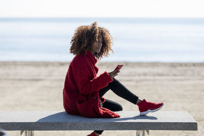 Woman using phone while sitting on bench at beach