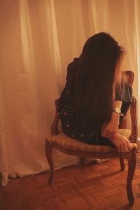 Woman with long hair sitting by curtain on chair at home