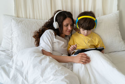 Family morning in bedroom. mom in headphones look at little kid son play games on tablet computer