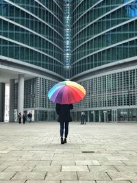 Rear view of woman under umbrella standing on footpath by modern building in city