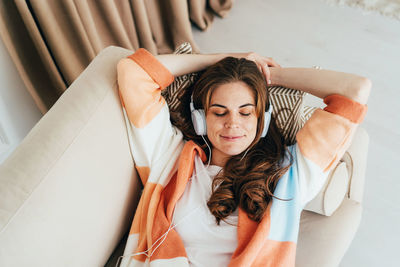 Young relaxed woman is laying down on sofa and listening to a music with headphones on her ears.