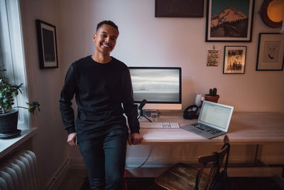 Portrait of smiling young freelance worker leaning on desk with computer at home