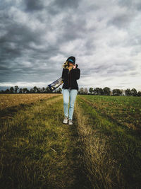 Full length of woman wearing baseball cap standing on farm against cloudy sky