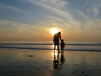 Silhouette rear view of man and son on wet shore at beach against sky during sunset