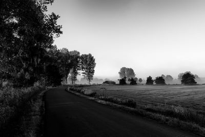 Empty road along trees on field against clear sky