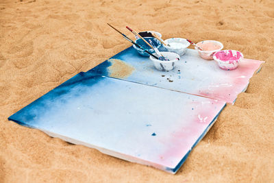 Two painted canvas in blue and pink colors with palettes of paints lying on sand, artist's palette