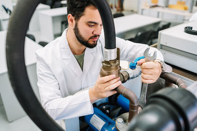 Focused male scientist in medical uniform tightening bolts with wrench on special machine with hose while working in modern light laboratory