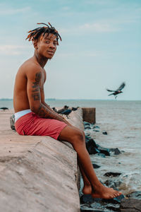 Full length of shirtless man sitting on concrete against sea and sky