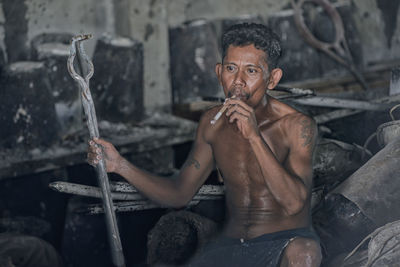 Shirtless man smoking cigar while sitting in workshop