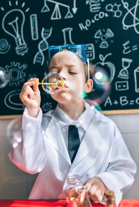 Boy wearing lab coat while blowing bubbles in classroom