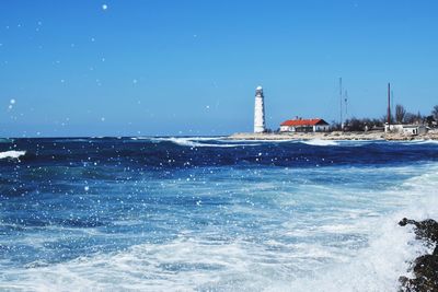 Lighthouse by sea against clear blue sky