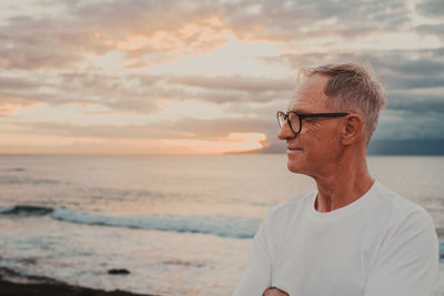 Portrait of young man standing at beach against sky during sunset