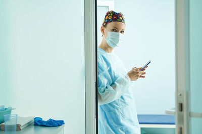 Side view of female doctor in medical uniform and sterile mask text messaging on cellphone in clinic