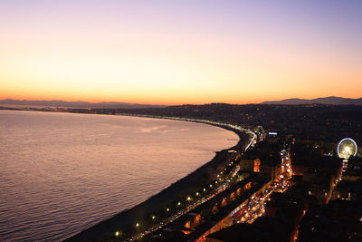 Aerial view of illuminated city by sea against orange sky during sunset