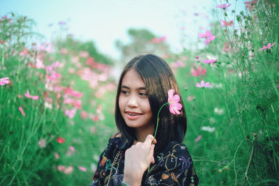 Smiling young woman holding flower while standing amidst plants