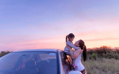 Woman holding girl on car against sky during sunset