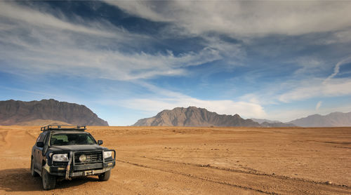 A rugged off-road vehicle travels through an expansive desert with majestic mountains and a blue sky