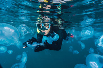 A woman in a black wetsuit is in the water with jellyfish. the jellyfish are floating around her