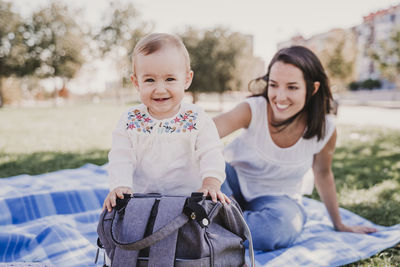 Mother looking at cute smiling baby daughter standing by backpack in park