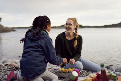 Smiling woman and daughter cooking corn on barbecue grill at beach during sunset