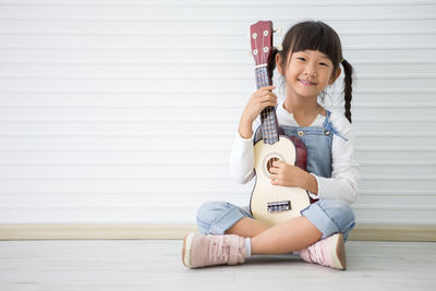 Full length portrait of cute girl playing ukelele against white wall at home 