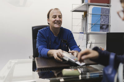 Positive adult male doctor in uniform giving payment terminal with plastic card to crop customer at table in veterinary