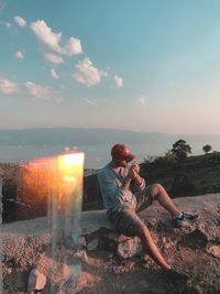 Man sitting on rock against sky during sunset